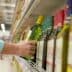 Individual reaching for a green bottle of olive oil on a supermarket shelf among other oil bottles. - Olive Oil Times