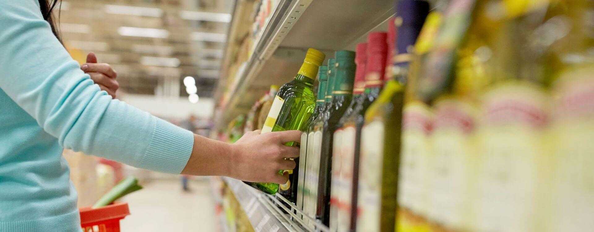 Individual reaching for a green bottle of olive oil on a supermarket shelf among other oil bottles. - Olive Oil Times