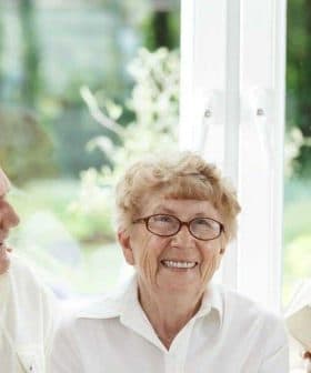 A group of seniors and a young woman sitting together, smiling and interacting indoors. - Olive Oil Times
