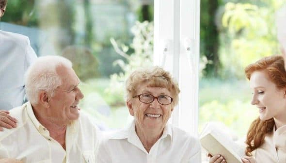 A group of seniors and a young woman sitting together, smiling and interacting indoors. - Olive Oil Times