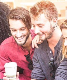 A group of six friends sitting together, looking at mobile devices and smiling. - Olive Oil Times