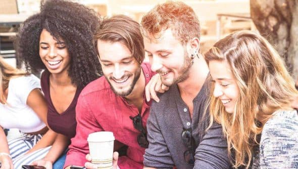 A group of six friends sitting together, looking at mobile devices and smiling. - Olive Oil Times
