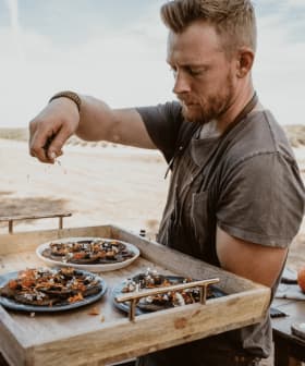 A man in a gray shirt garnishing plates of food on a wooden tray outdoors. - Olive Oil Times