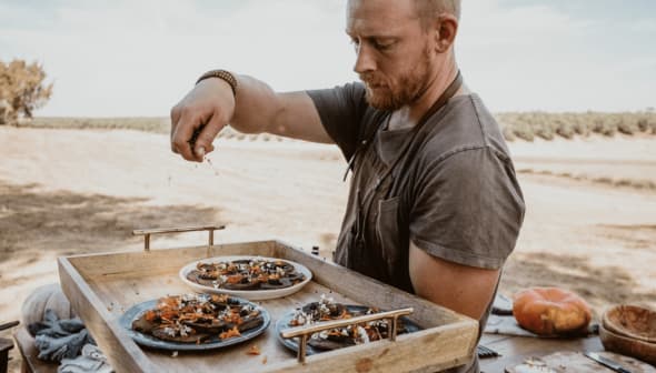 A man in a gray shirt garnishing plates of food on a wooden tray outdoors. - Olive Oil Times