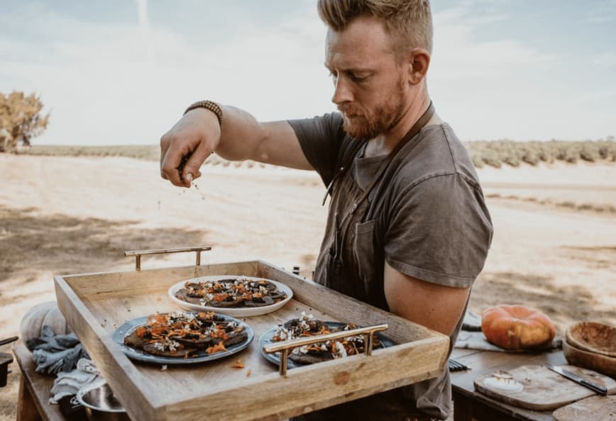 A man in a gray shirt garnishing plates of food on a wooden tray outdoors. - Olive Oil Times
