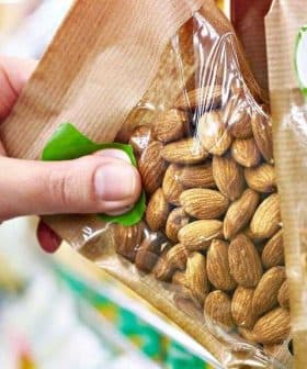 A hand reaching for a transparent package of almonds on a store shelf. - Olive Oil Times