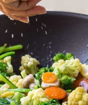 Person sprinkling salt over a mix of vegetables in a black wok with a wooden spoon. - Olive Oil Times