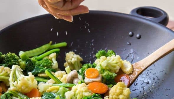 Person sprinkling salt over a mix of vegetables in a black wok with a wooden spoon. - Olive Oil Times