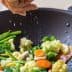Person sprinkling salt over a mix of vegetables in a black wok with a wooden spoon. - Olive Oil Times