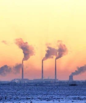 Three smoke stacks emitting smoke against a colorful sunset sky with a snowy landscape in the foreground. - Olive Oil Times
