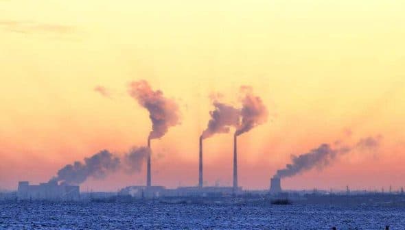 Three smoke stacks emitting smoke against a colorful sunset sky with a snowy landscape in the foreground. - Olive Oil Times