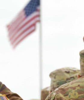 Two military personnel in camouflage uniforms saluting the American flag during a ceremony. - Olive Oil Times