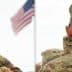 Two military personnel in camouflage uniforms saluting the American flag during a ceremony. - Olive Oil Times