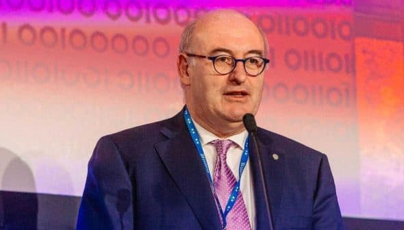 Phil Hogan speaking at a podium during an event with a microphone in front of a colorful background. - Olive Oil Times