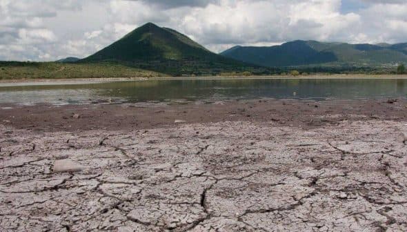 Dry, cracked earth with a mountain in the background under a cloudy sky. - Olive Oil Times