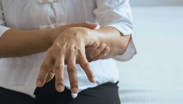 Individual with a white shirt holding their wrist while seated on a bed. - Olive Oil Times