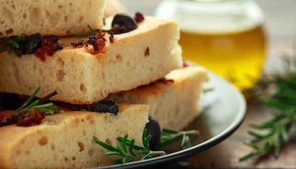 Slices of focaccia bread topped with olives and herbs on a black plate. - Olive Oil Times