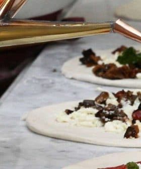 A copper oil dispenser pouring extra virgin olive oil onto a pizza base with toppings. - Olive Oil Times