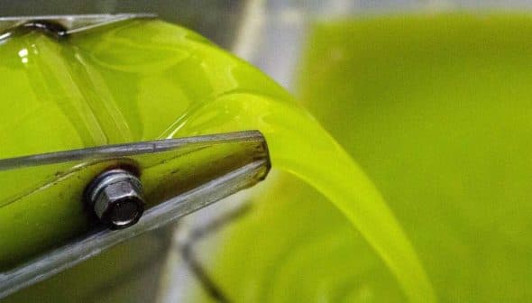 Stream of green olive oil flowing from a machine into a container during the extraction process. - Olive Oil Times