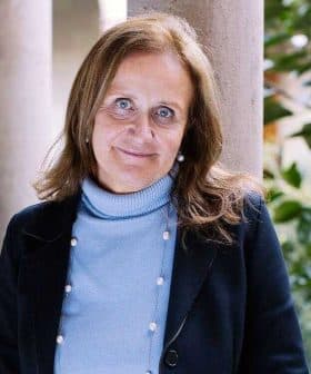 Woman with brown hair wearing a blue sweater and black jacket standing in a corridor with columns. - Olive Oil Times