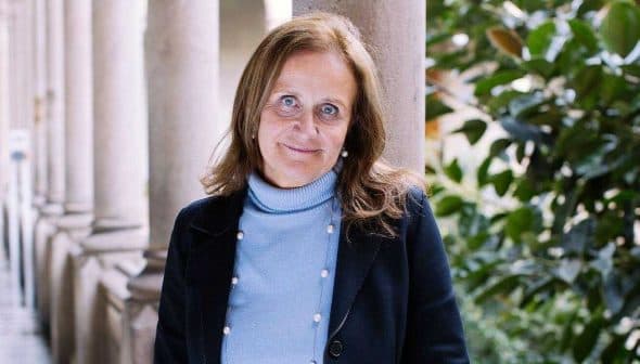 Woman with brown hair wearing a blue sweater and black jacket standing in a corridor with columns. - Olive Oil Times