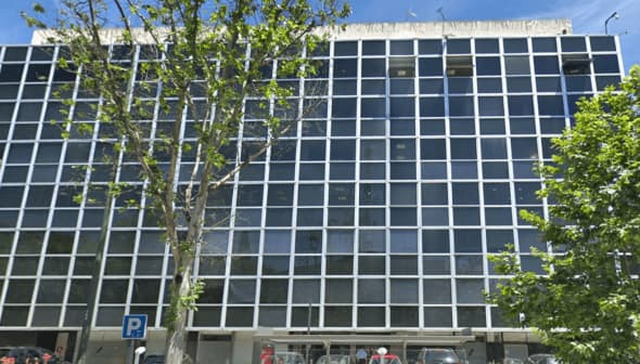 A modern office building featuring a grid pattern of large windows and a tree in front. - Olive Oil Times