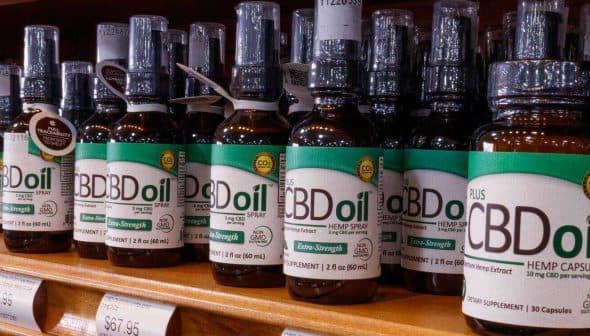 Row of CBD oil bottles with various labels displayed on a wooden shelf in a store. - Olive Oil Times
