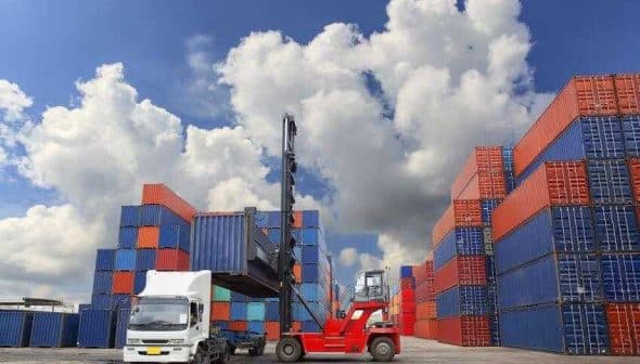 A forklift lifting a shipping container in a storage area surrounded by stacked containers. - Olive Oil Times