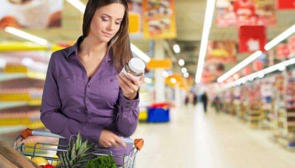 Woman examining a product label while holding a jar in a grocery store aisle. - Olive Oil Times