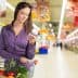 Woman examining a product label while holding a jar in a grocery store aisle. - Olive Oil Times