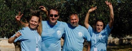 Four individuals wearing blue t-shirts posing together outdoors under olive trees. - Olive Oil Times