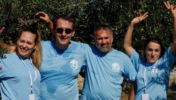 Four individuals wearing blue t-shirts posing together outdoors under olive trees. - Olive Oil Times