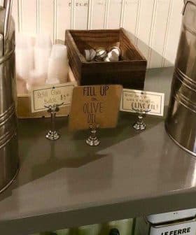 Two stainless steel dispensers labeled for olive oil and vinegar on a table with utensils. - Olive Oil Times