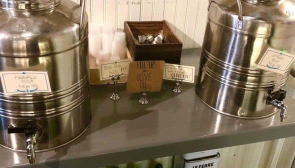 Two stainless steel dispensers labeled for olive oil and vinegar on a table with utensils. - Olive Oil Times