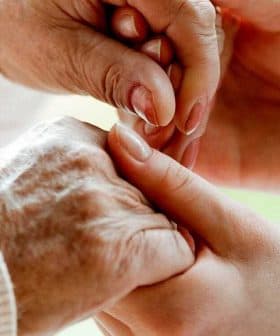 An elderly person's hand being held by a caregiver's hand in a supportive gesture. - Olive Oil Times