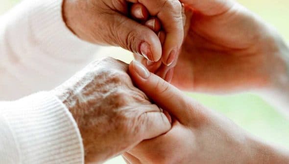 An elderly person's hand being held by a caregiver's hand in a supportive gesture. - Olive Oil Times
