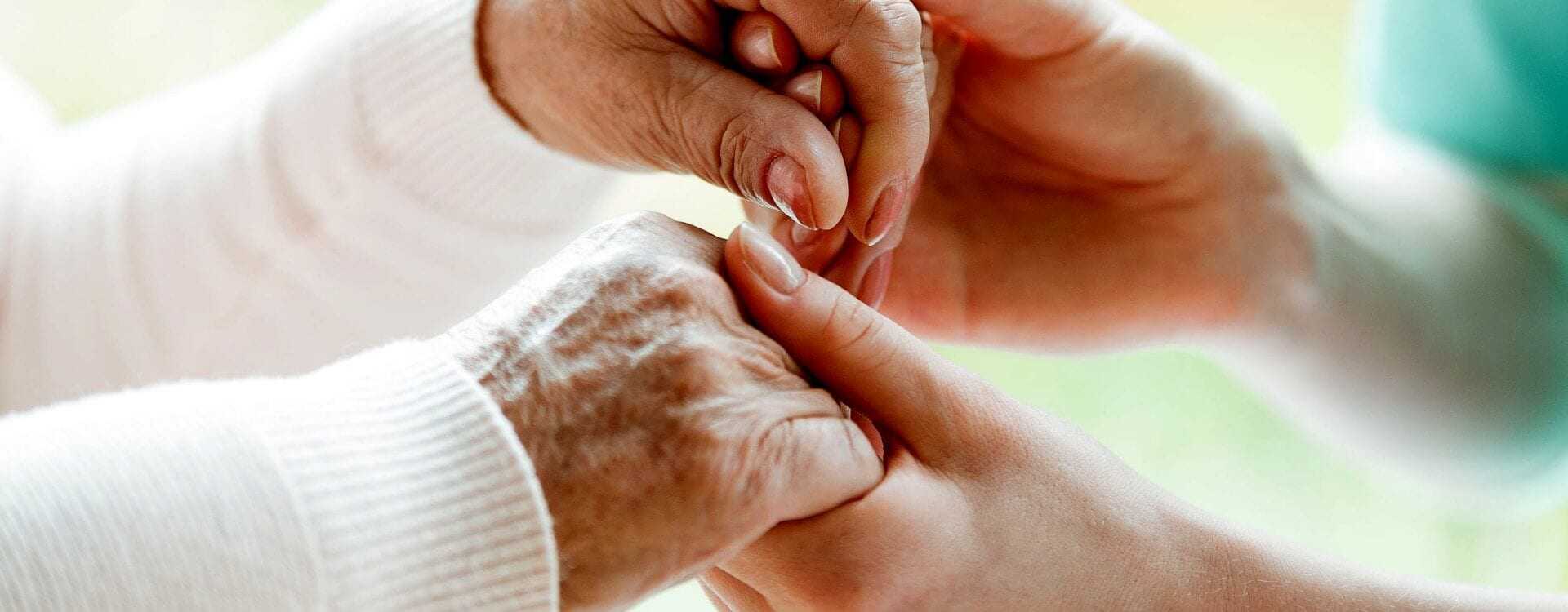 An elderly person's hand being held by a caregiver's hand in a supportive gesture. - Olive Oil Times