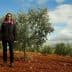 Individual standing in an olive grove with green trees and red soil in the background. - Olive Oil Times
