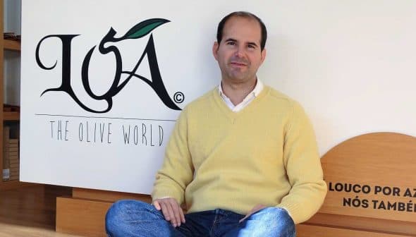 Man wearing a yellow sweater sitting on the floor in front of the LOA logo. - Olive Oil Times