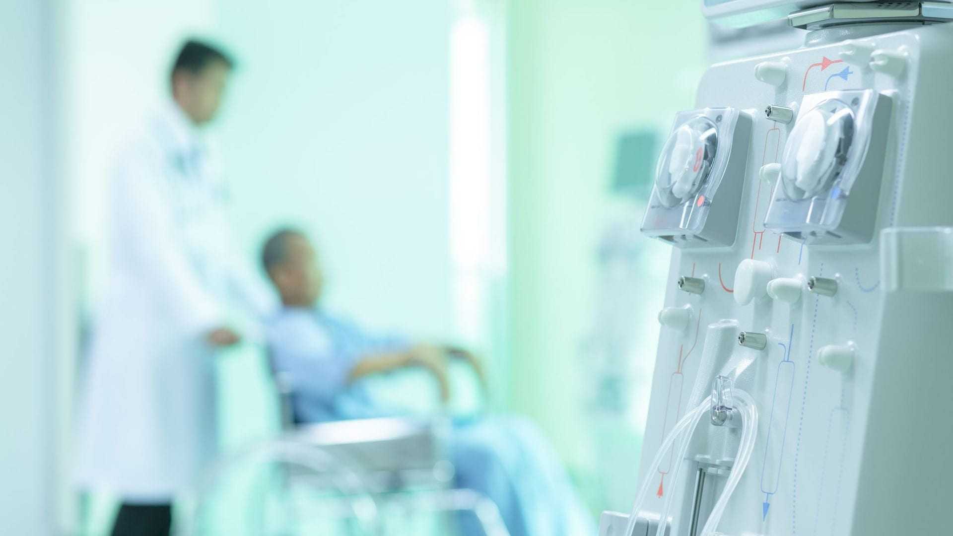 Dialysis machine with dials and tubes in a medical environment with a patient in a wheelchair in the background. - Olive Oil Times
