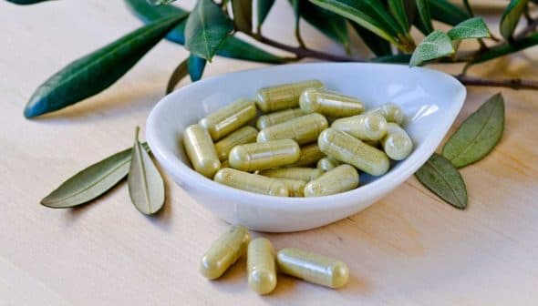 A white dish containing green herbal capsules, surrounded by leaves on a wooden surface. - Olive Oil Times
