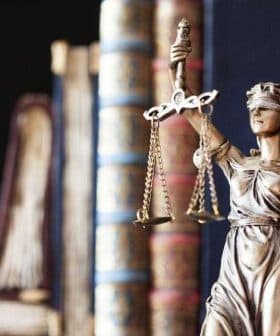 Bronze statue of Lady Justice holding scales and wearing a blindfold, with books in the background. - Olive Oil Times