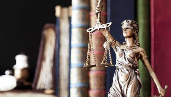 Bronze statue of Lady Justice holding scales and wearing a blindfold, with books in the background. - Olive Oil Times