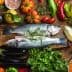 Two whole fish placed on a wooden board surrounded by various fresh vegetables and herbs. - Olive Oil Times