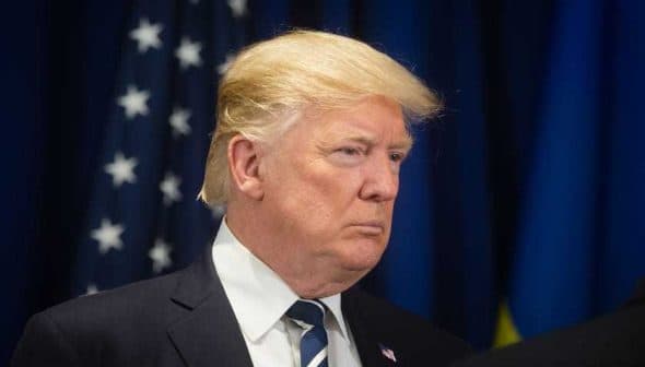 Man with blonde hair wearing a suit and tie, facing to the side against a dark background with flags. - Olive Oil Times