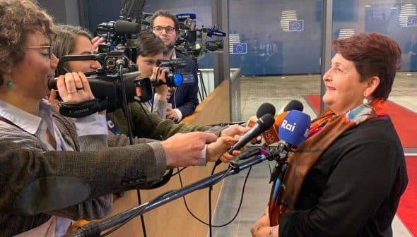 A woman speaking to reporters while being filmed by multiple cameras in a press setting. - Olive Oil Times