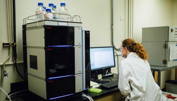 Laboratory technician in a white coat operating analytical equipment and reviewing data on a computer screen. - Olive Oil Times