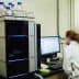 Laboratory technician in a white coat operating analytical equipment and reviewing data on a computer screen. - Olive Oil Times