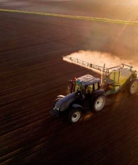 A black tractor equipped with a sprayer applying pesticides on a brown agricultural field during sunset. - Olive Oil Times