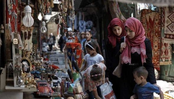 Two women wearing hijabs walking through a market with children and various items for sale. - Olive Oil Times